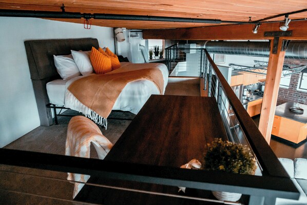 A modern loft-style bedroom with a large bed, a wooden dining table, and a view of the kitchen and living area.