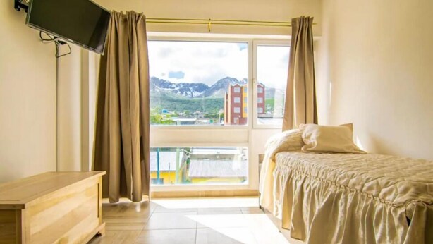 A hotel room with a bed, a TV, a wooden dresser, and a balcony with a mountain view. - Mirador de los Cerros (Ushuaia)