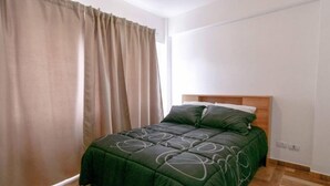 A single bed with a dark green bedspread and matching pillows, a wooden headboard, and a window with beige curtains. - Mirador de los Cerros (Ushuaia)