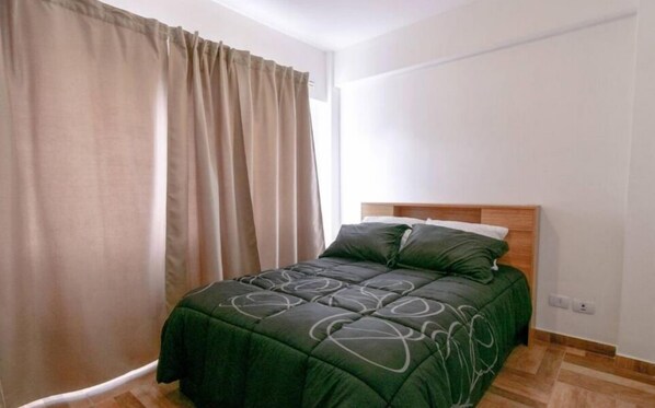 A single bed with a dark green bedspread and matching pillows, a wooden headboard, and a window with beige curtains.