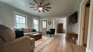A spacious living room with a wooden floor, a ceiling fan, a sofa, a coffee table, and a TV mounted on the wall.
