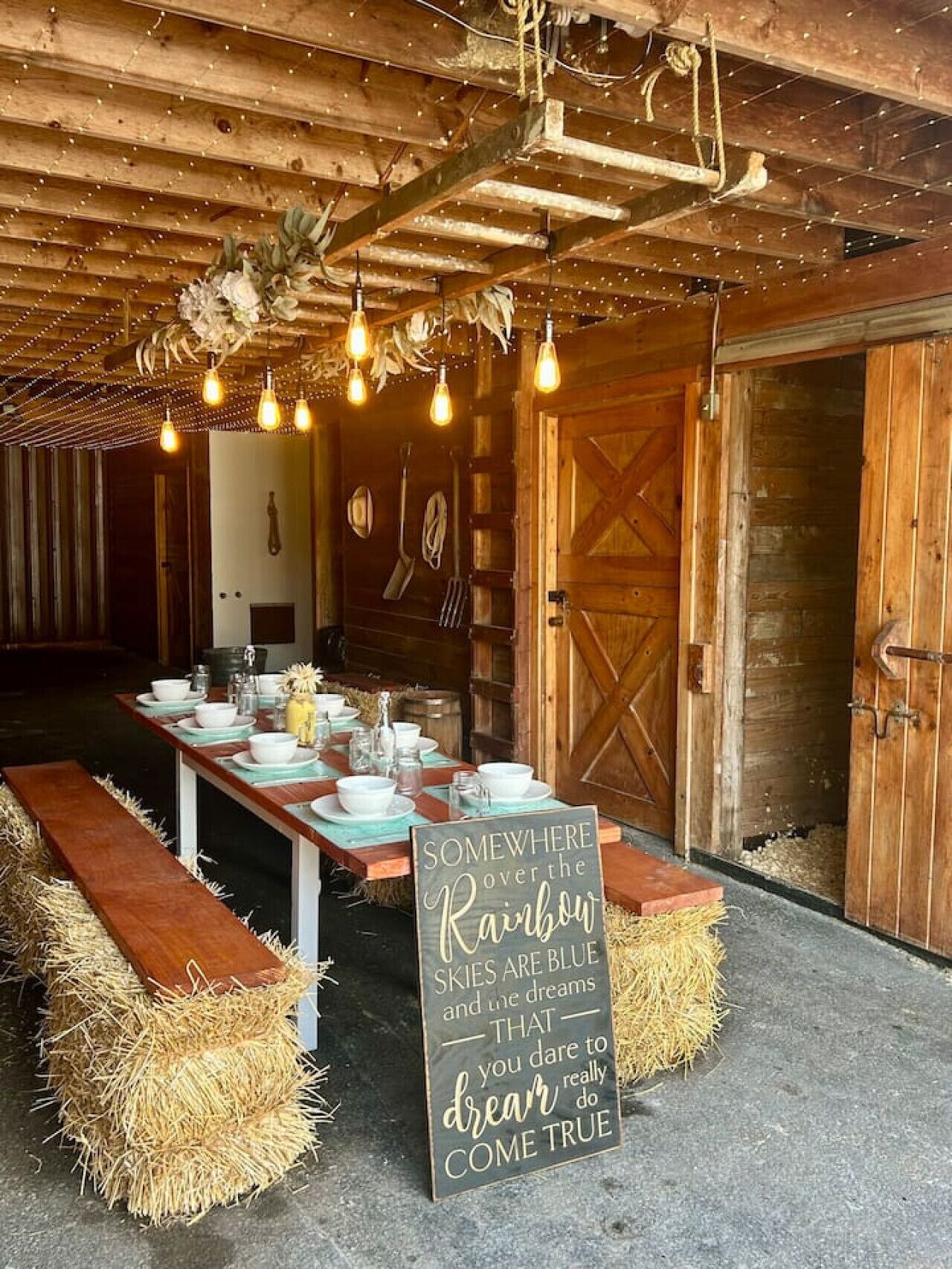 A rustic dining area with a long table set for a meal, hay bales on either side, and a sign with a motivational quote.