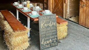 A rustic dining area with a long table set for a meal, hay bales on either side, and a sign with a motivational quote.