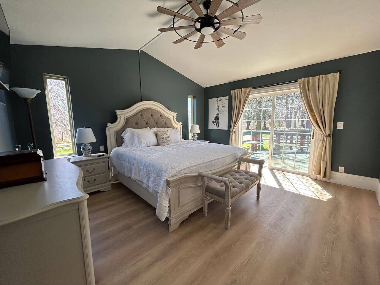 Quarto com uma cama grande, um ventilador de teto, uma mesa de cabeceira, uma luminária e uma janela com cortinas.