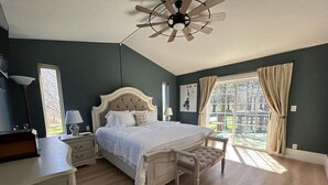 A bedroom with a large bed, a ceiling fan, a nightstand, a lamp, and a window with curtains.