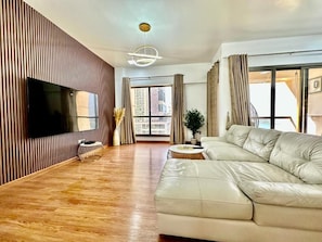 A modern living room with a large sofa, a flat-screen TV mounted on a wooden wall, a potted plant, and a view of the city through large windows. - 3BR Luxury Apartment Sea View (Dubai)