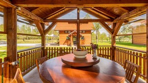 A wooden gazebo with a round table and chairs, overlooking a wooden house and a grassy area.