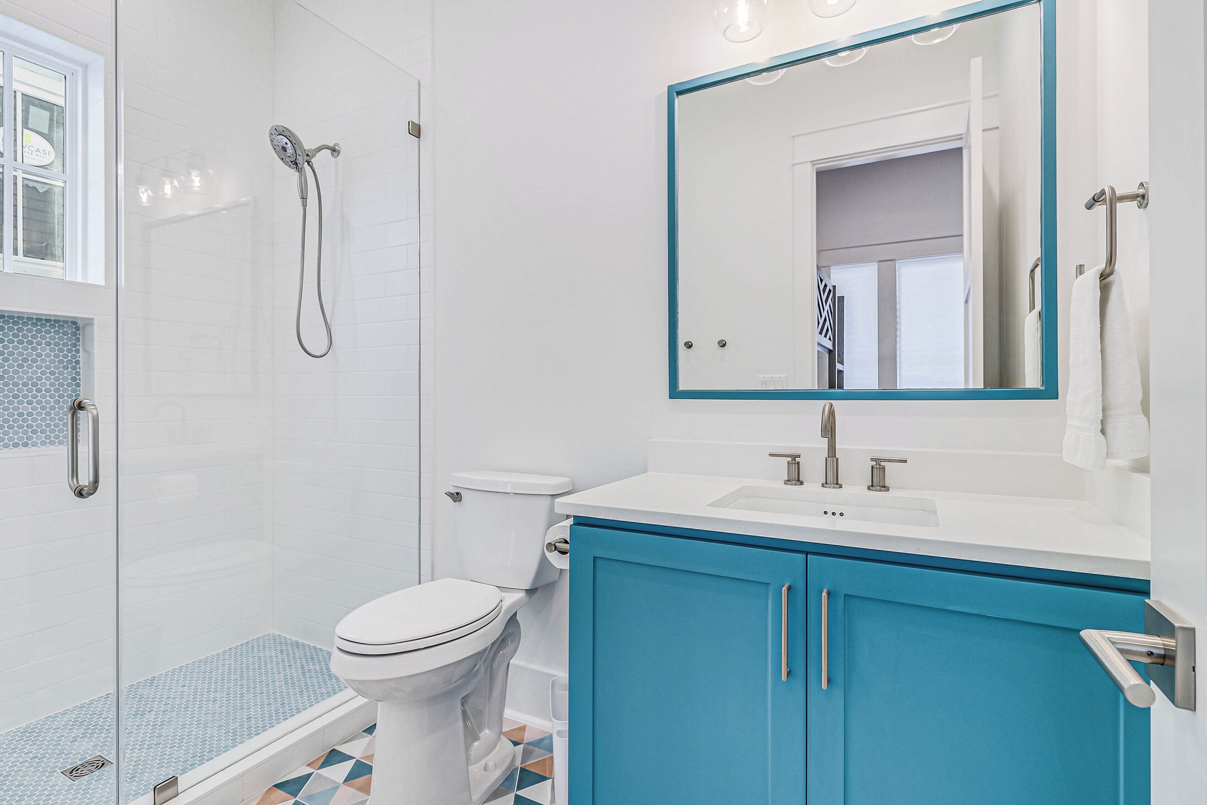 A modern bathroom with a glass shower enclosure, a white toilet, a sink with a blue cabinet, and a mirror with a teal frame.