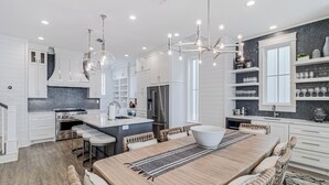 A modern kitchen with white cabinetry, a central island, and a dining area with a wooden table and bar stools. - Sea Vous Play 112 NEI (Port Aransas)