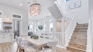 Une salle à manger spacieuse, dotée d’une grande table en bois et de six chaises, d’un lustre et d’un escalier avec des rampes blanches.