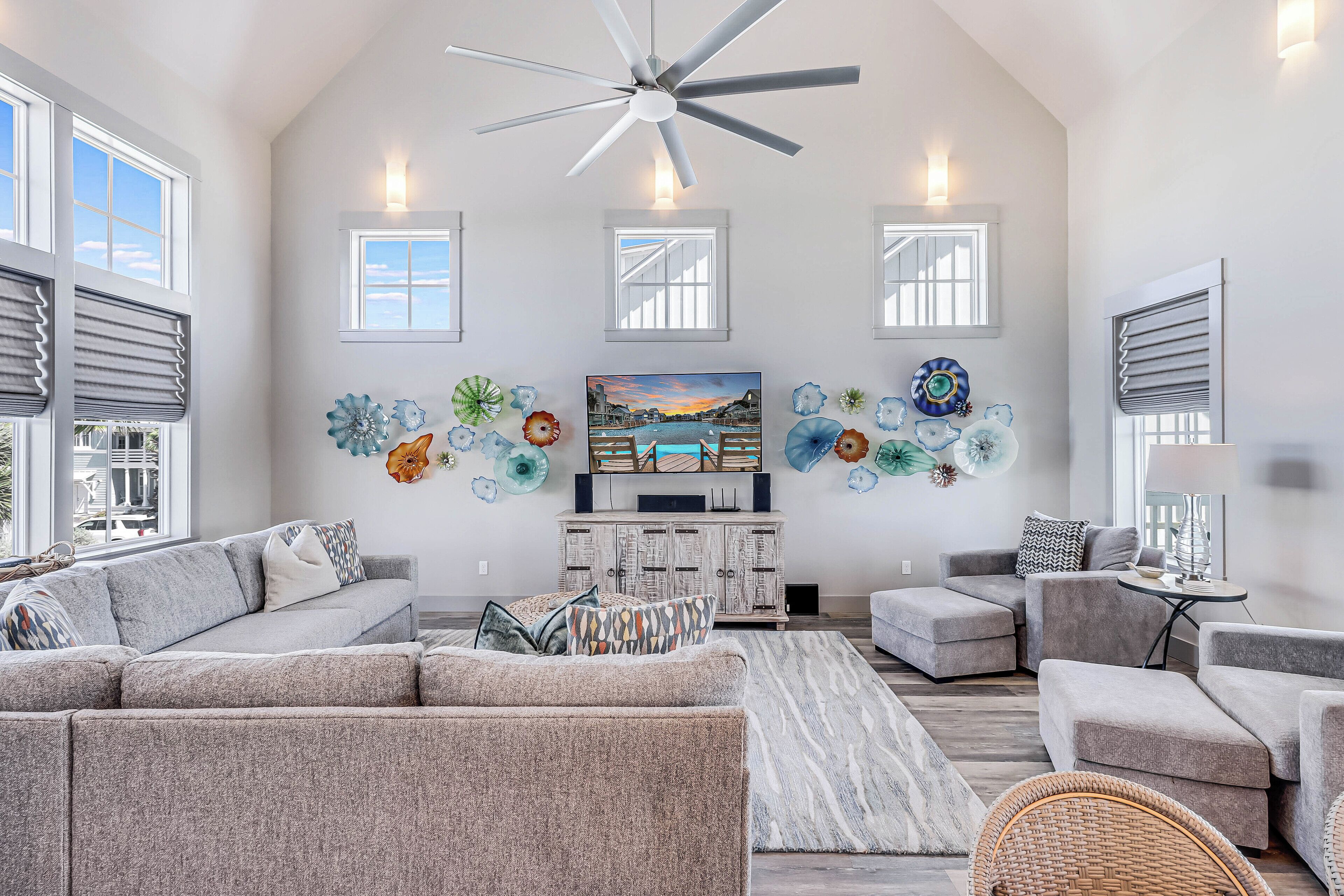 A spacious living room with a high ceiling, large windows, and a ceiling fan.