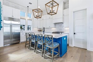 Uma cozinha moderna com armários brancos, uma geladeira de aço inoxidável, uma ilha azul com banquetas e luminárias pendentes geométricas.