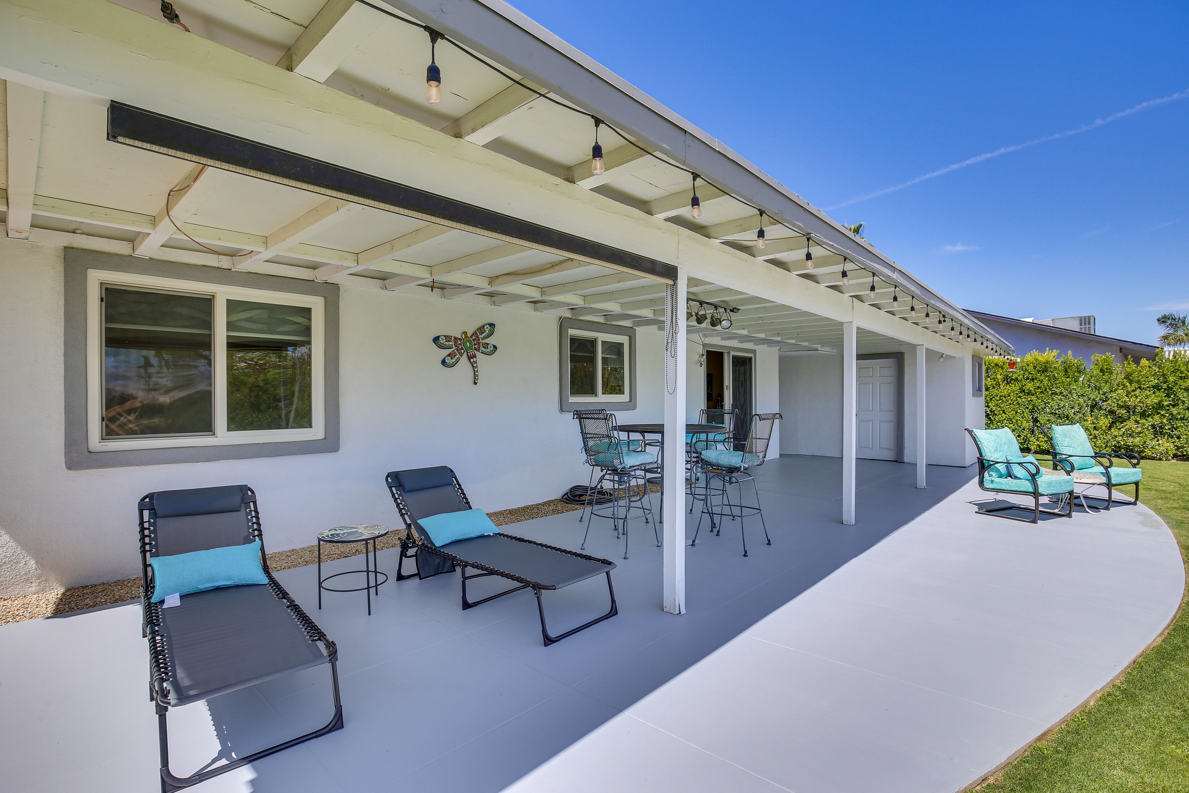 A covered patio area with lounge chairs and a table, a white exterior wall, and a clear blue sky.