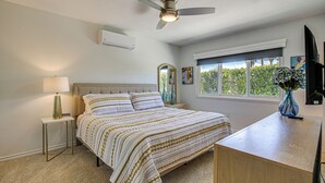 Ein Schlafzimmer mit Bett, Nachttisch, Lampe, Deckenventilator, Klimaanlage, Fenster und Kunstwerk.