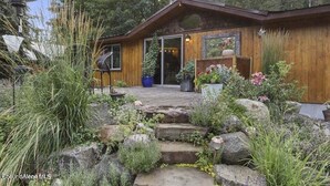 A wooden cabin with a stone pathway leading to the entrance, surrounded by lush vegetation and a large window overlooking the garden. - Priest Lake - Escape to the Lake (Nordman)