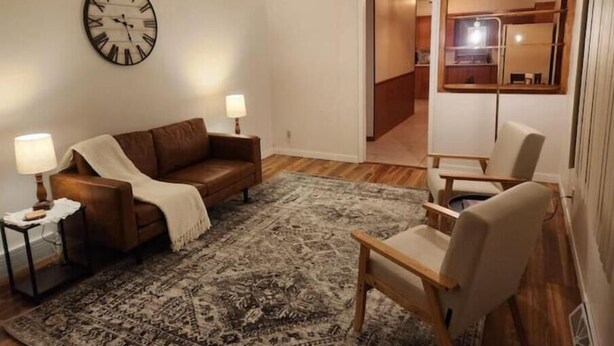A living room with a brown leather sofa, a patterned rug, a wooden chair, and a wall clock. - Hill Cottage (Park River)