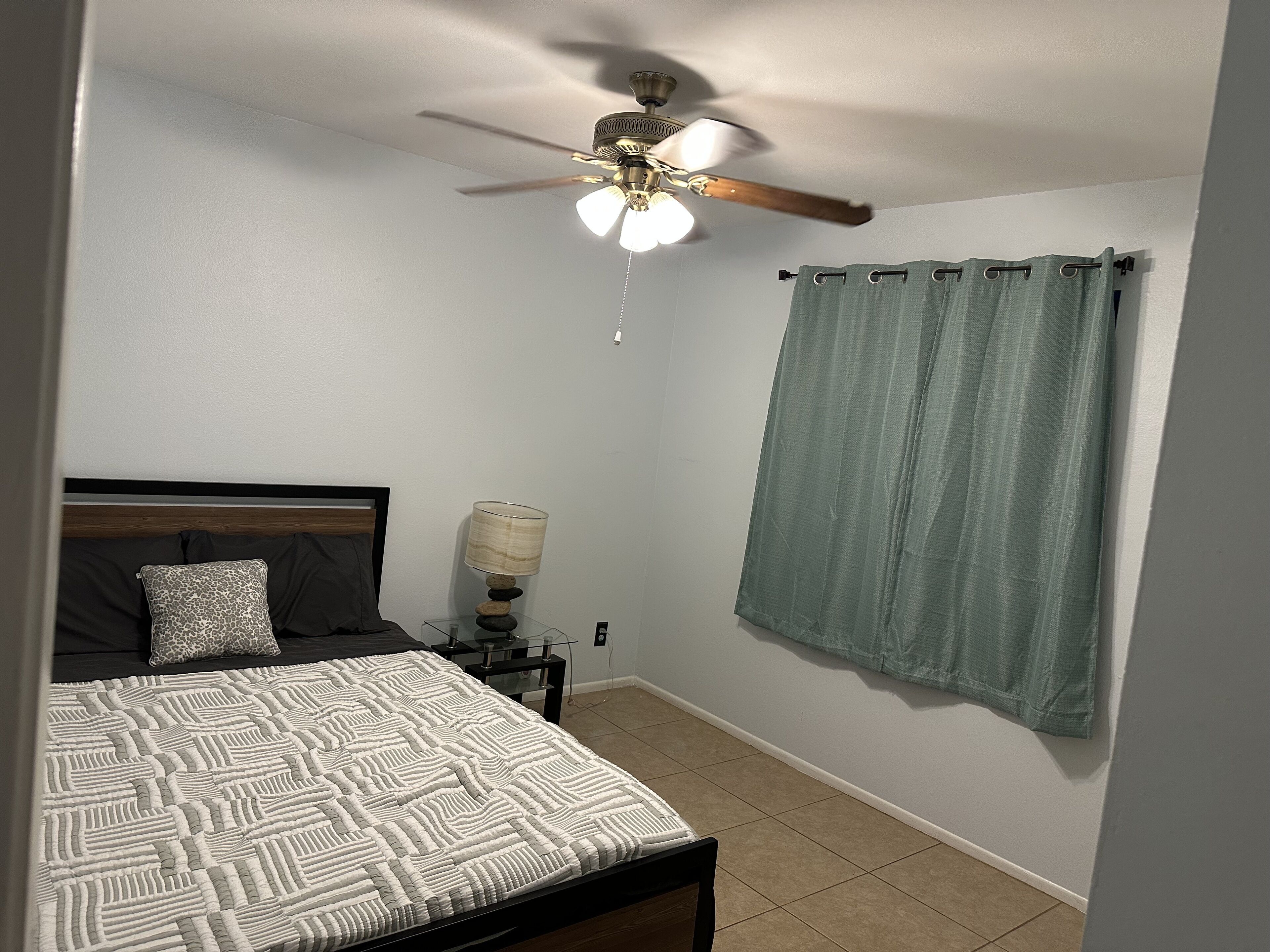 A bedroom with a bed, a nightstand, a lamp, a ceiling fan, and a curtain.
