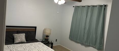 A bedroom with a bed, a nightstand, a lamp, a ceiling fan, and a curtain.