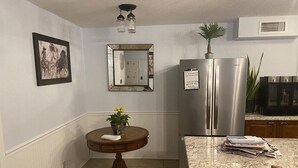 A kitchen with a stainless steel refrigerator, a round wooden table with a potted plant, and a marble countertop with papers.