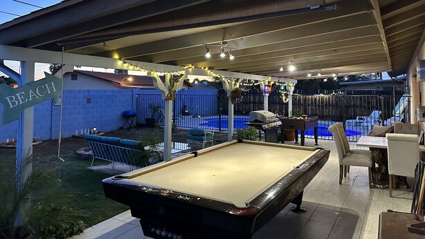 A pool table in a covered outdoor area with a 'BEACH' sign, a pool, and outdoor seating.