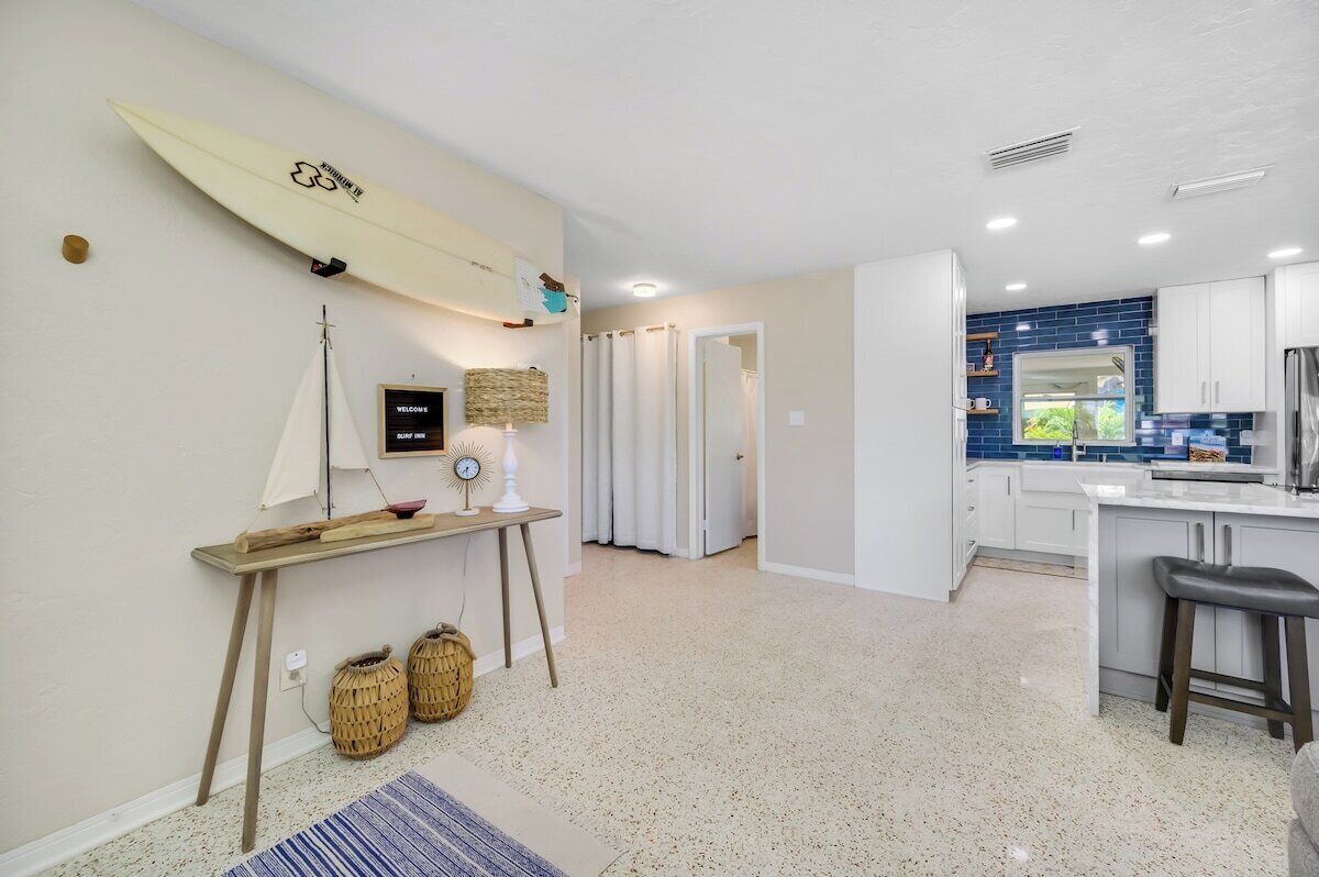 A modern kitchen with white cabinets and a blue backsplash, a dining area with a bar stool, and a surfboard decoration on the wall.