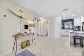 A modern kitchen with white cabinets and a blue backsplash, a dining area with a bar stool, and a surfboard decoration on the wall.