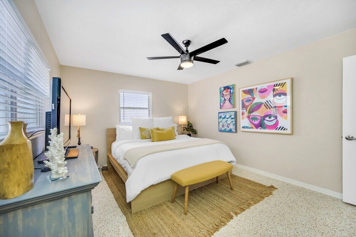A bedroom with a bed, bedside tables, a ceiling fan, artwork on the wall, and a window with blinds.