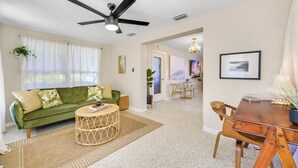 Uma sala de estar com sofá verde, uma mesa de centro redonda, uma mesa de jantar com cadeiras, um ventilador de teto e um candelabro.