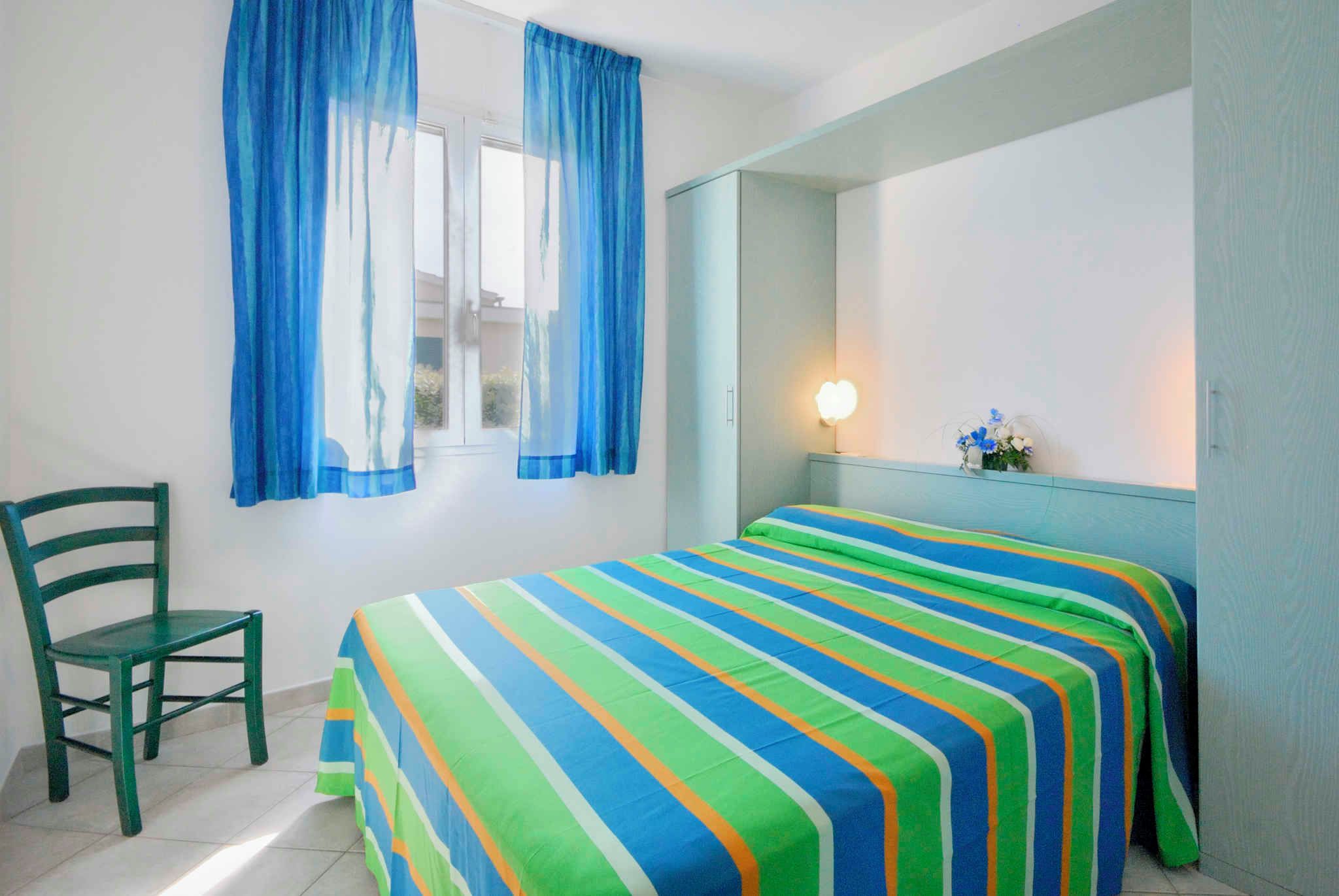Ein Zimmer mit einem Bett, einem Sessel, einem Fenster mit Vorhängen, einer Nachttischlampe und einem Spiegel an der Wand.