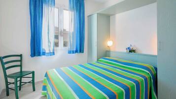 Ein Zimmer mit einem Bett, einem Sessel, einem Fenster mit Vorhängen, einer Nachttischlampe und einem Spiegel an der Wand.