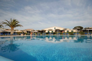 Ein Swimmingpool mit blauen Stühlen und Sonnenschirmen, umgeben von Gebäuden bei blauem Himmel.