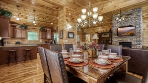 Un interior de madera con una mesa de comedor para cuatro, una chimenea de piedra y una zona de cocina con armarios de madera y taburetes de bar.