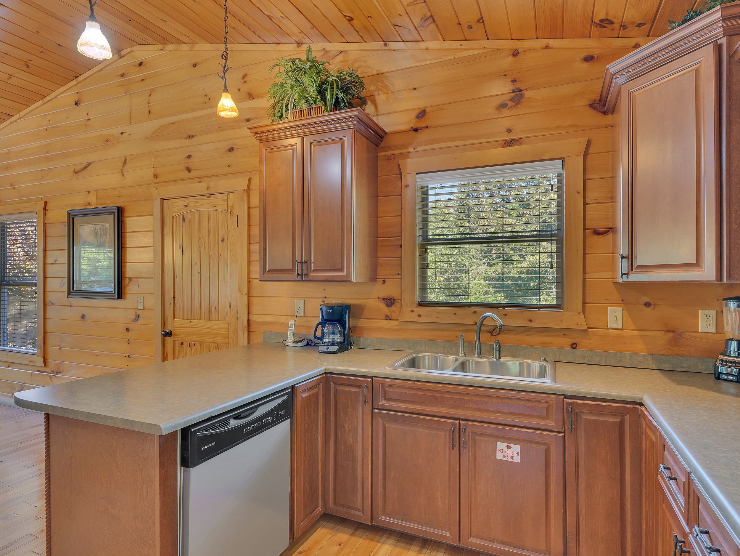 A wooden cabin kitchen with a built-in dishwasher, a coffee maker, and a window with a view of trees.