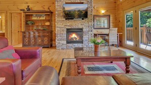 Interior acogedor de una cabaña con chimenea de piedra, paredes de madera, un televisor montado arriba y una zona de estar cómoda con sofás de cuero y una mesita de café.