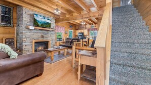 A cozy cabin interior with a stone fireplace, wooden furniture, a staircase, and a view of the outdoors. - ER81 - Family Making Memories (Pigeon Forge)