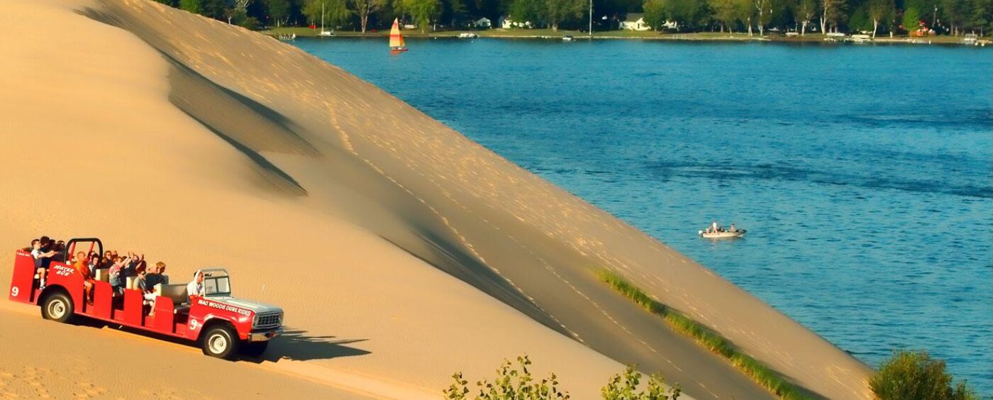 A dune buggy tour vehicle with passengers, a sandy dune, and a body of water with a sailboat in the background.
