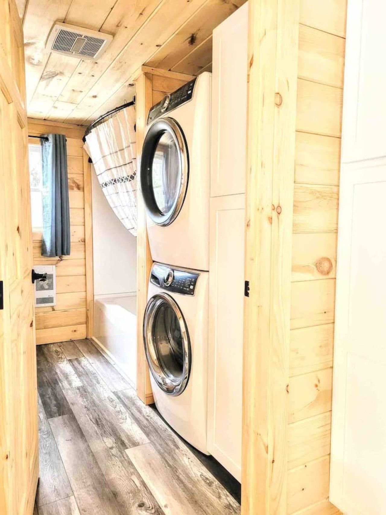 A laundry room with two washing machines, wooden walls, and a window with curtains.