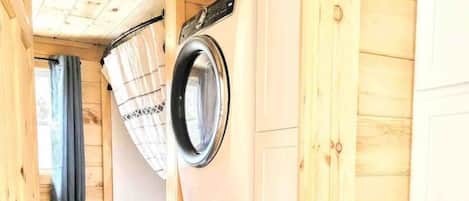A laundry room with two washing machines, wooden walls, and a window with curtains.