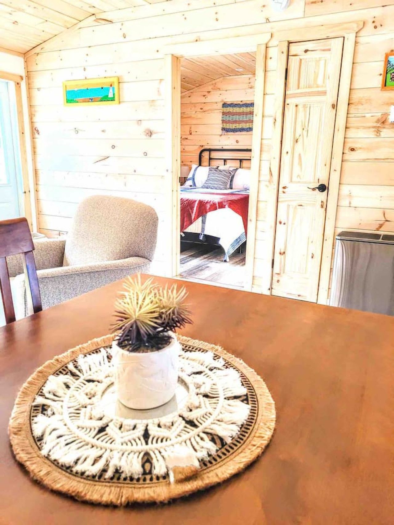 A wooden cabin interior with a dining table, a chair, a bed with a red bedspread, and a painting on the wall.