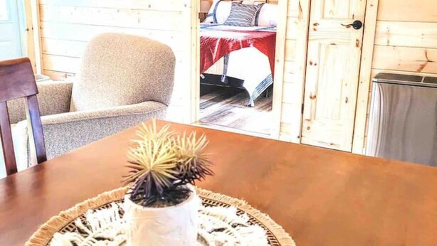Een houten chalet van binnen met een eettafel, een stoel, een bed met een rode deken en een schilderij aan de muur.