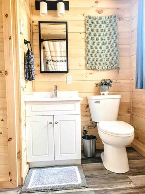 Une salle de bain rustique, avec des murs en bois, comprenant un lavabo blanc, un miroir, des toilettes et une petite poubelle.