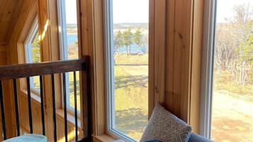 Ein gemütliches Blockhauss-Interieur mit einem großen Fenster, das Blick auf einen See und Bäume bietet.
