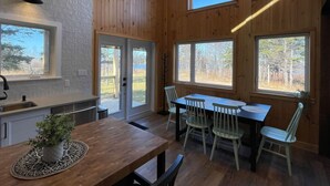 A wooden interior with a dining table and chairs, a kitchen area with a sink, and large windows offering views of the outdoors. - Chalet Gedouque, a tranquille retreat located on the Cabot Trail with waterfront views (Chéticamp)