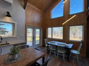 Un interior de madera con una mesa de comedor y sillas, una zona de cocina con un lavabo, y ventanas grandes que ofrecen vistas al exterior.