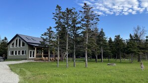 A two-story house with a sloped roof, a porch with wooden railings, and a grassy yard with trees. - Chalet Gedouque, a tranquille retreat located on the Cabot Trail with waterfront views (Chéticamp)
