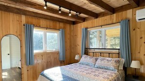 A wooden-paneled bedroom with a bed, two windows with curtains, a mirror, and a nightstand. - Chalet Gedouque, a tranquille retreat located on the Cabot Trail with waterfront views (Chéticamp)