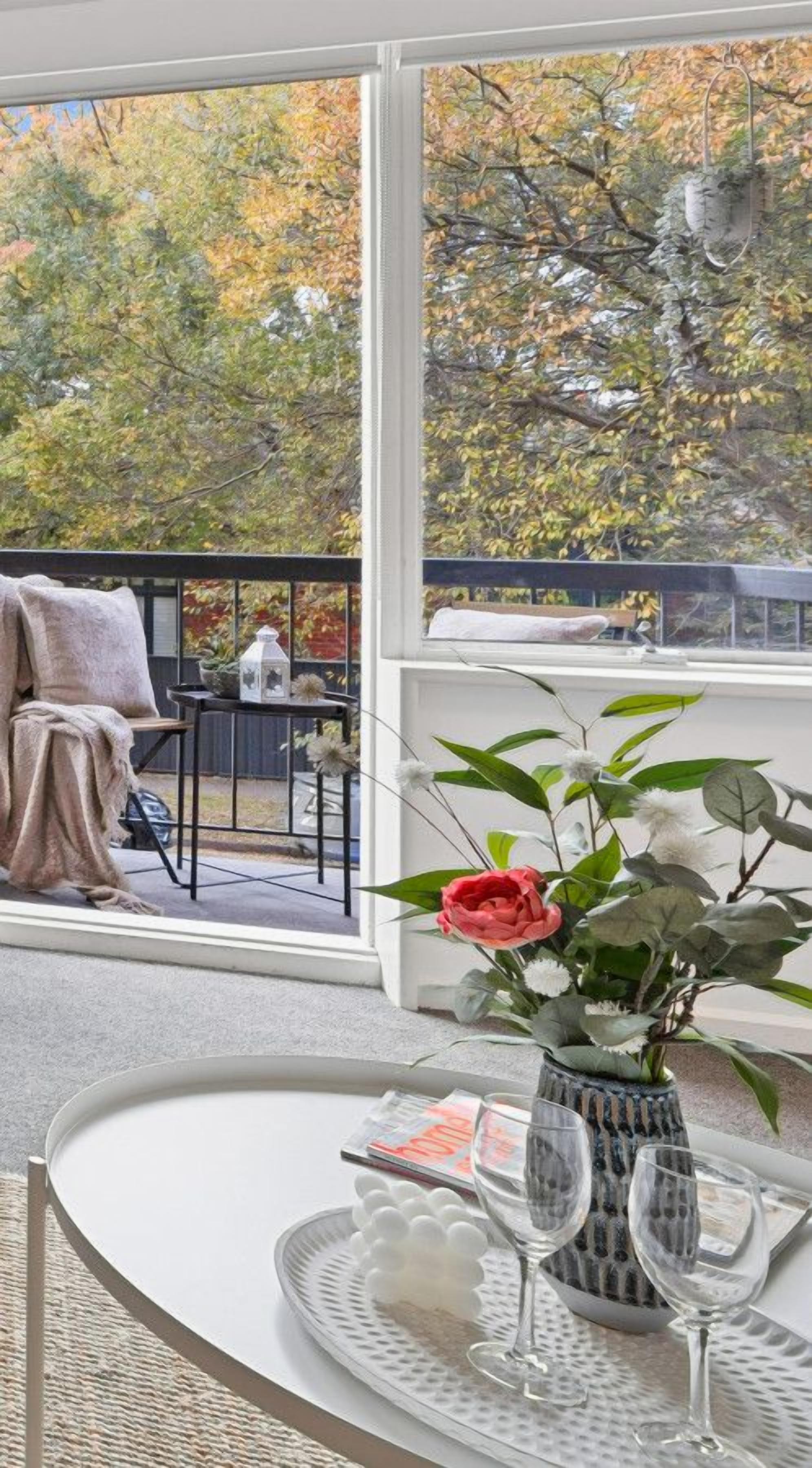 Ein Zimmer mit einem großen Fenster, einem Tisch mit einer Vase und Gläsern sowie einem Stuhl mit einer Decke.