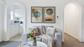 A modern living room with a grey sofa, a white coffee table, and a vase with flowers. There are two framed abstract paintings on the wall, and a kitchen is visible in the background.