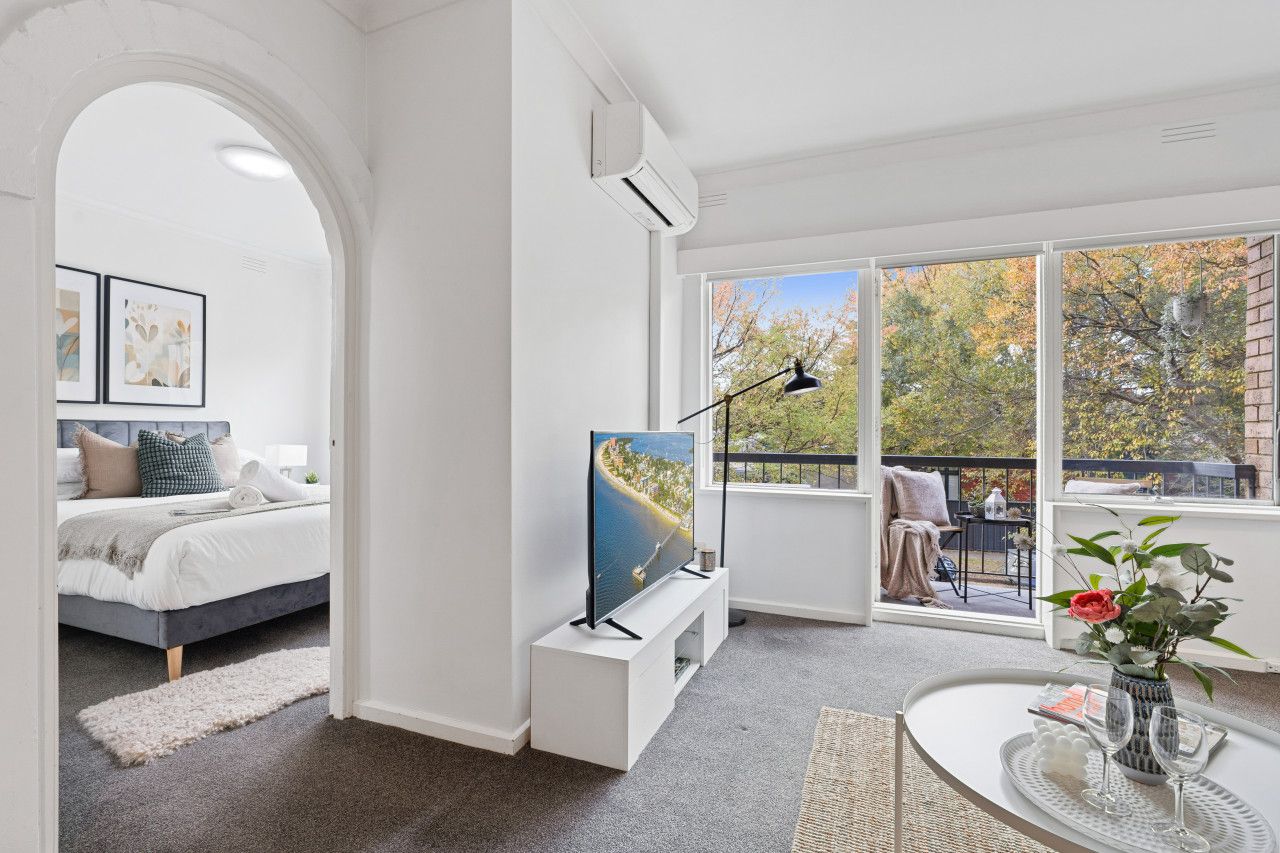 Ein modernes Schlafzimmer mit Bett, Fernseher und Balkon mit Blick auf Bäume und Gebäude.