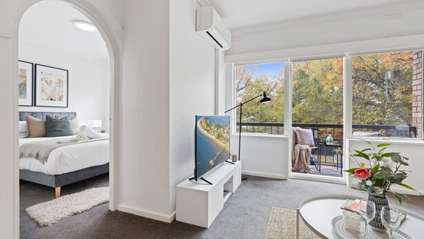Una camera da letto moderna con un letto, una TV e un balcone vista alberi e edifici.